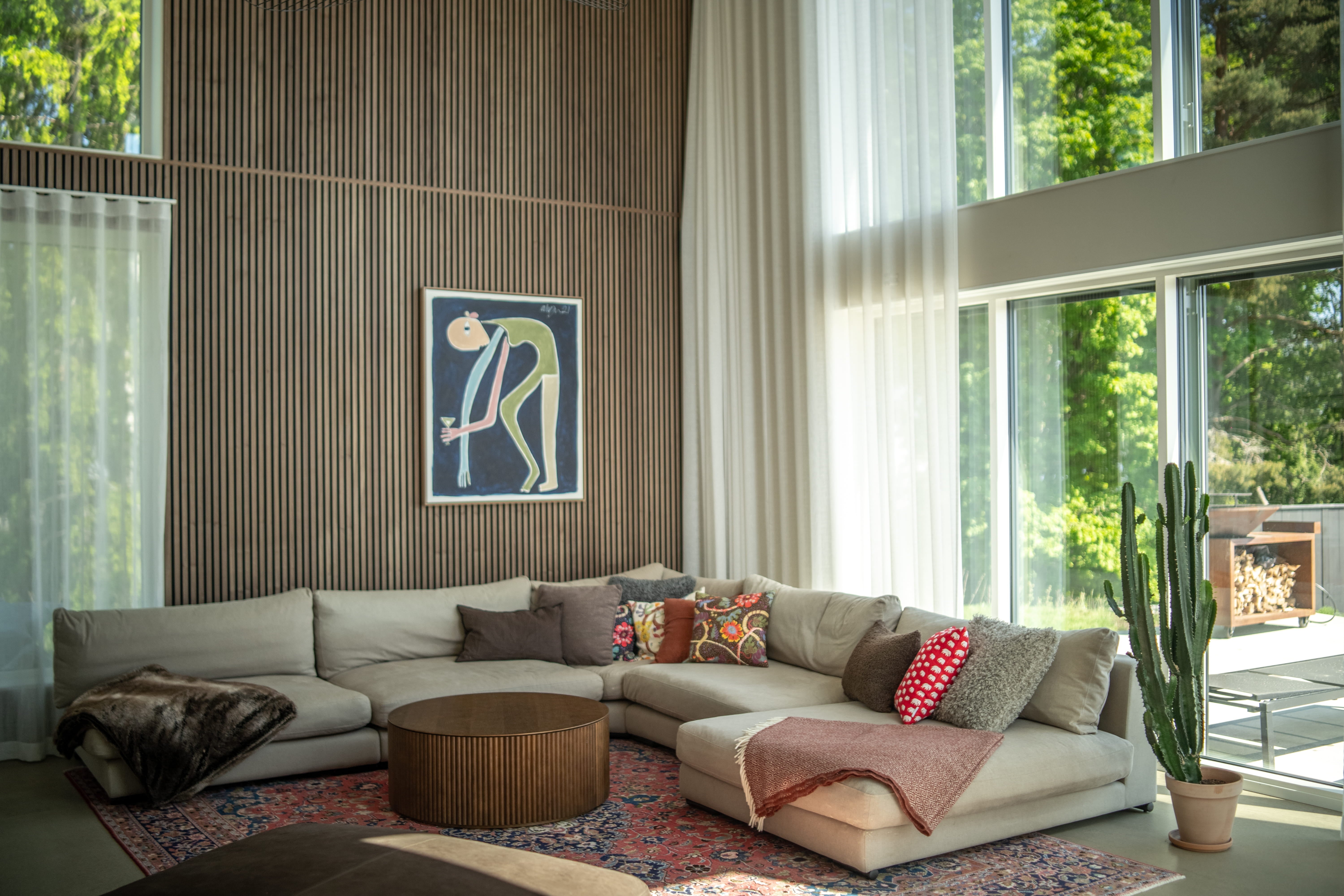 Modern living room with a large beige sectional sofa, colorful pillows, round coffee table, cactus, and floor-to-ceiling windows with sheer curtains and Nordic acoustic panels.