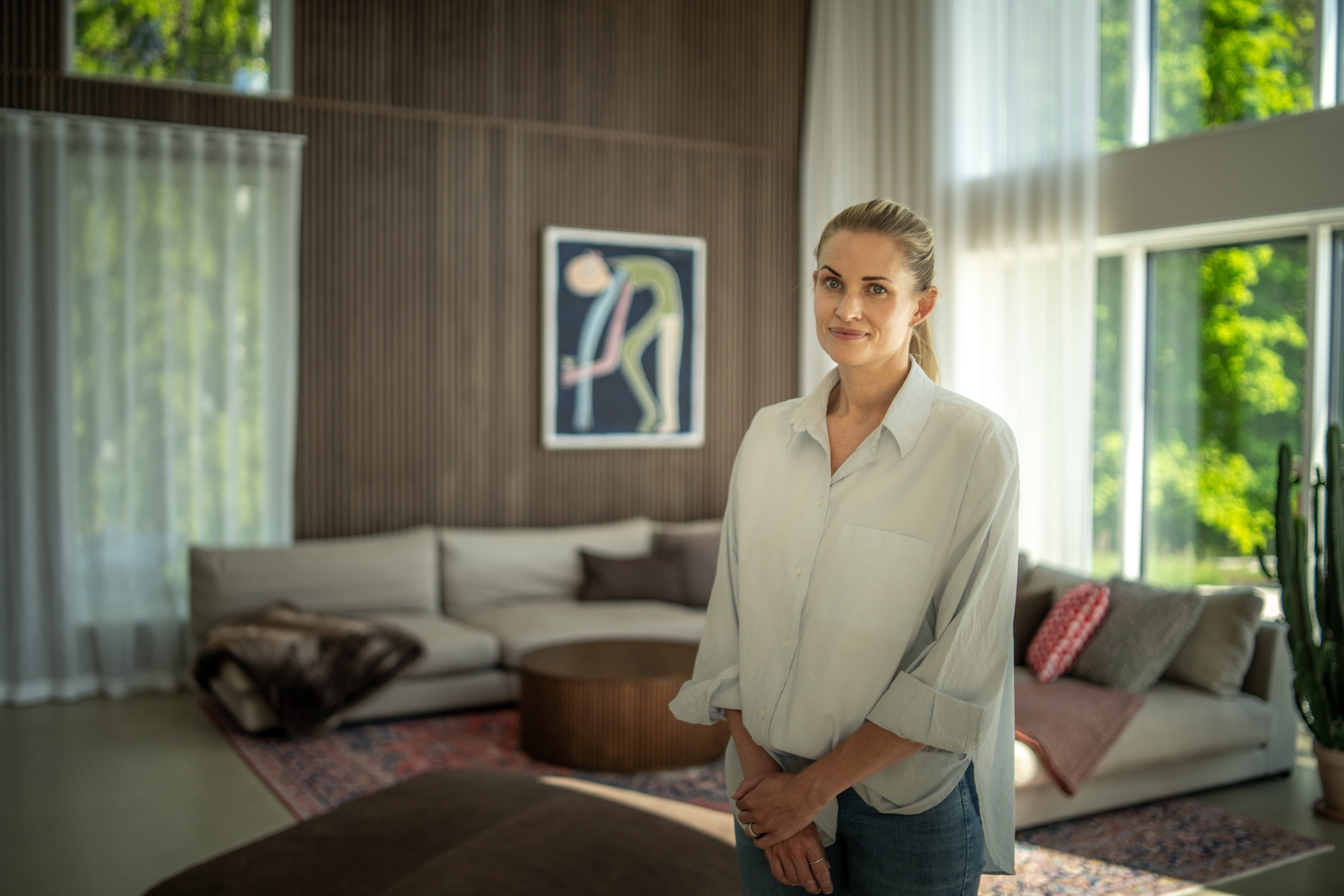 A woman stands in a sunlit living room with large windows, Nordic acoustic wood panels on the wall, contemporary art, and neutral-toned minimalist furniture.