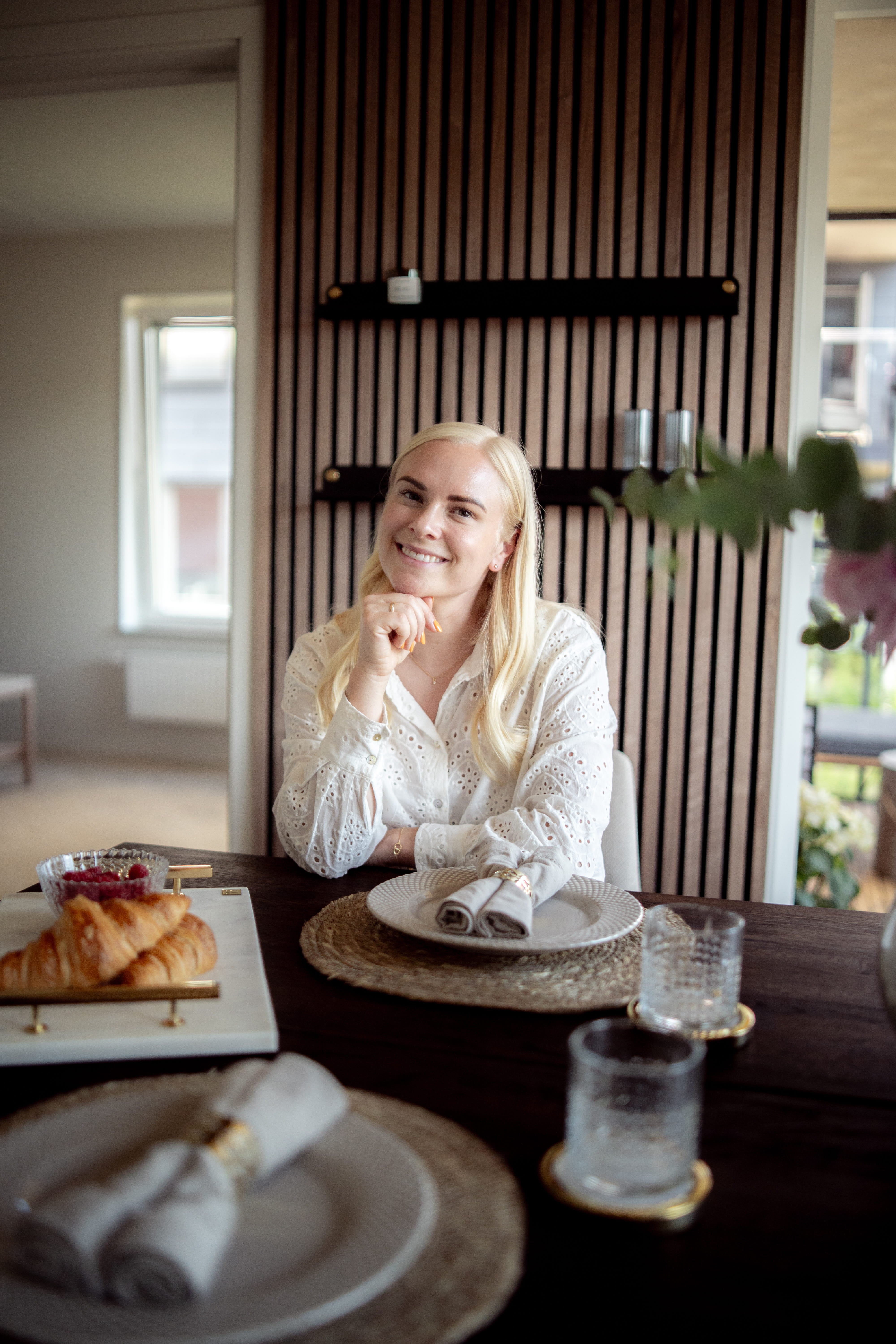 A woman with long blonde hair sits at a dining table set for breakfast with croissants and glassware, next to a Nordic acoustic panel on the wall.