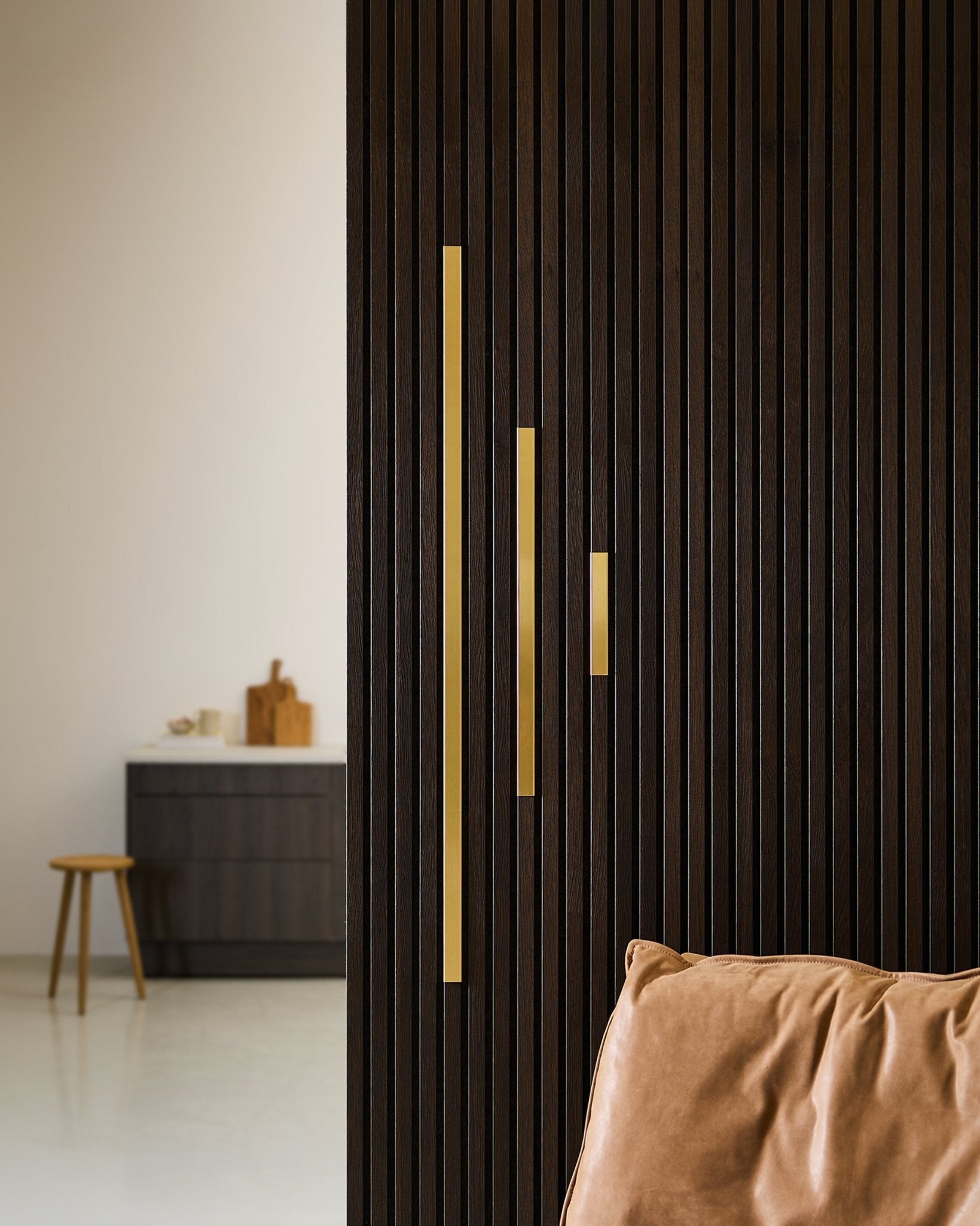 Modern interior with a WoodUpp brass-accented acoustic slatwall panel, gold handles, tan cushion, wooden stool, and dark cabinet.