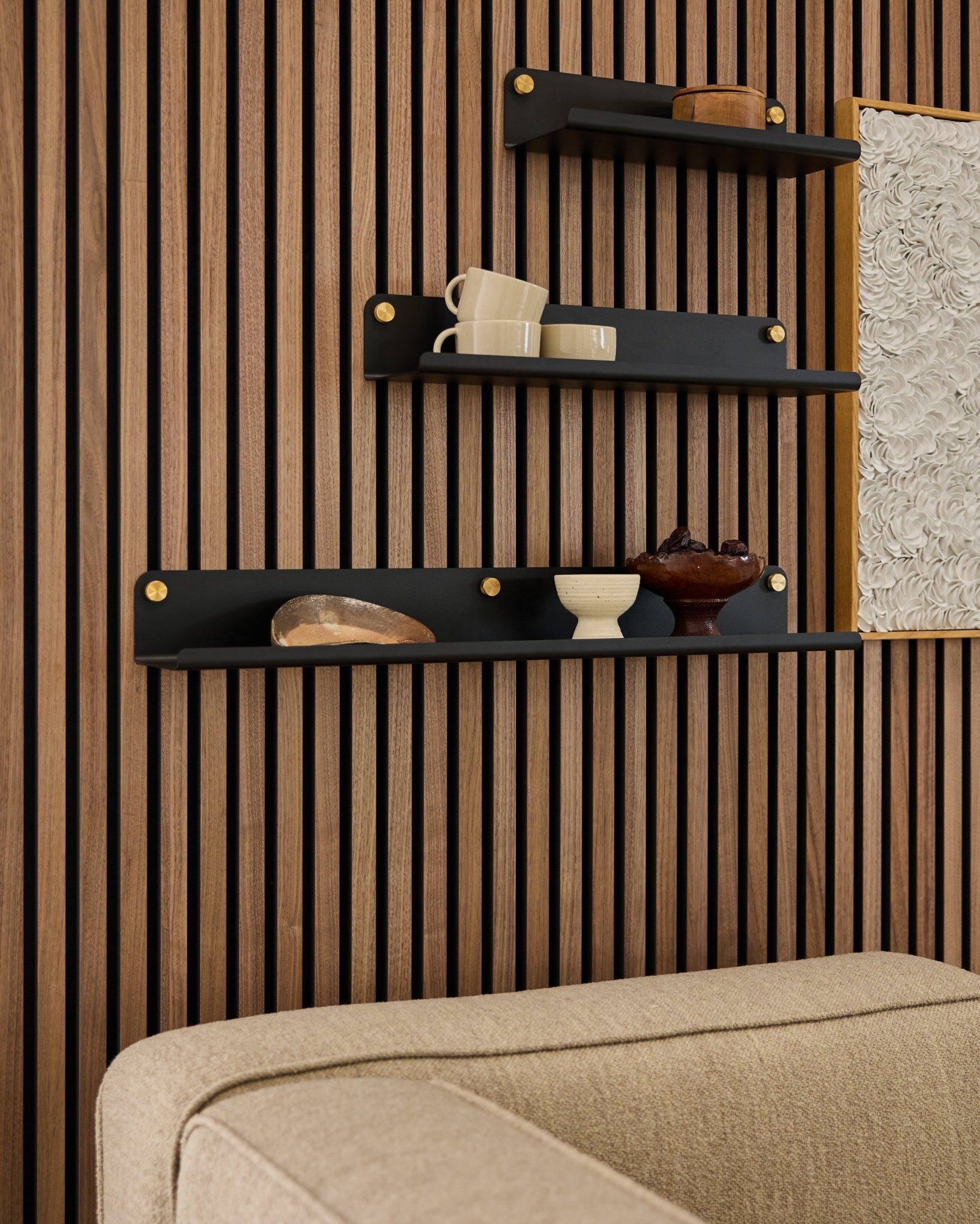 Three black shelves, including the WoodUpp Norr Wide Shelf (75.9 cm), display decor on a wood slat acoustic panel wall above a beige sofa; partial artwork visible.