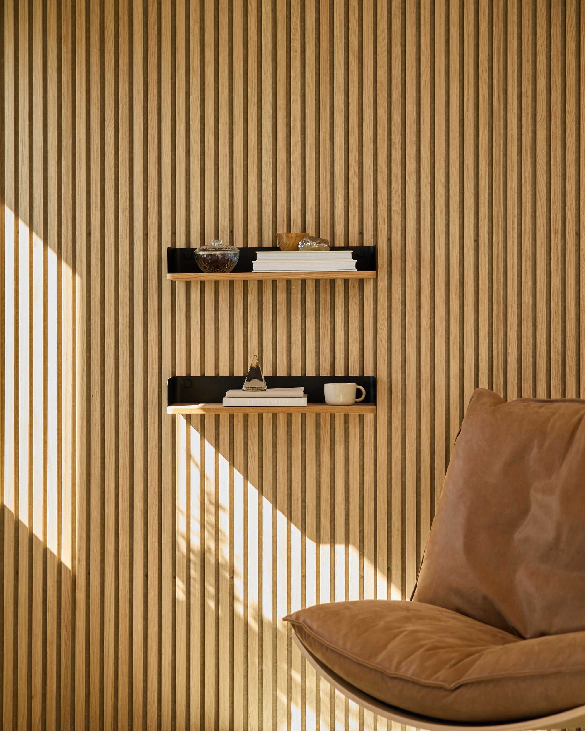 Modern Nordic acoustic wood panel wall with minimalist shelves holding books and decor, next to a tan leather chair illuminated by sunlight.