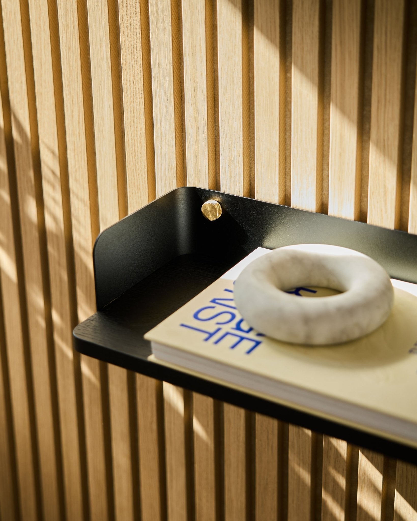 Akupanel Black Oak bookshelf by WoodUpp, 55.9 cm, holds a cream book with blue text and a round gray stone in front of vertical acoustic wood slats.