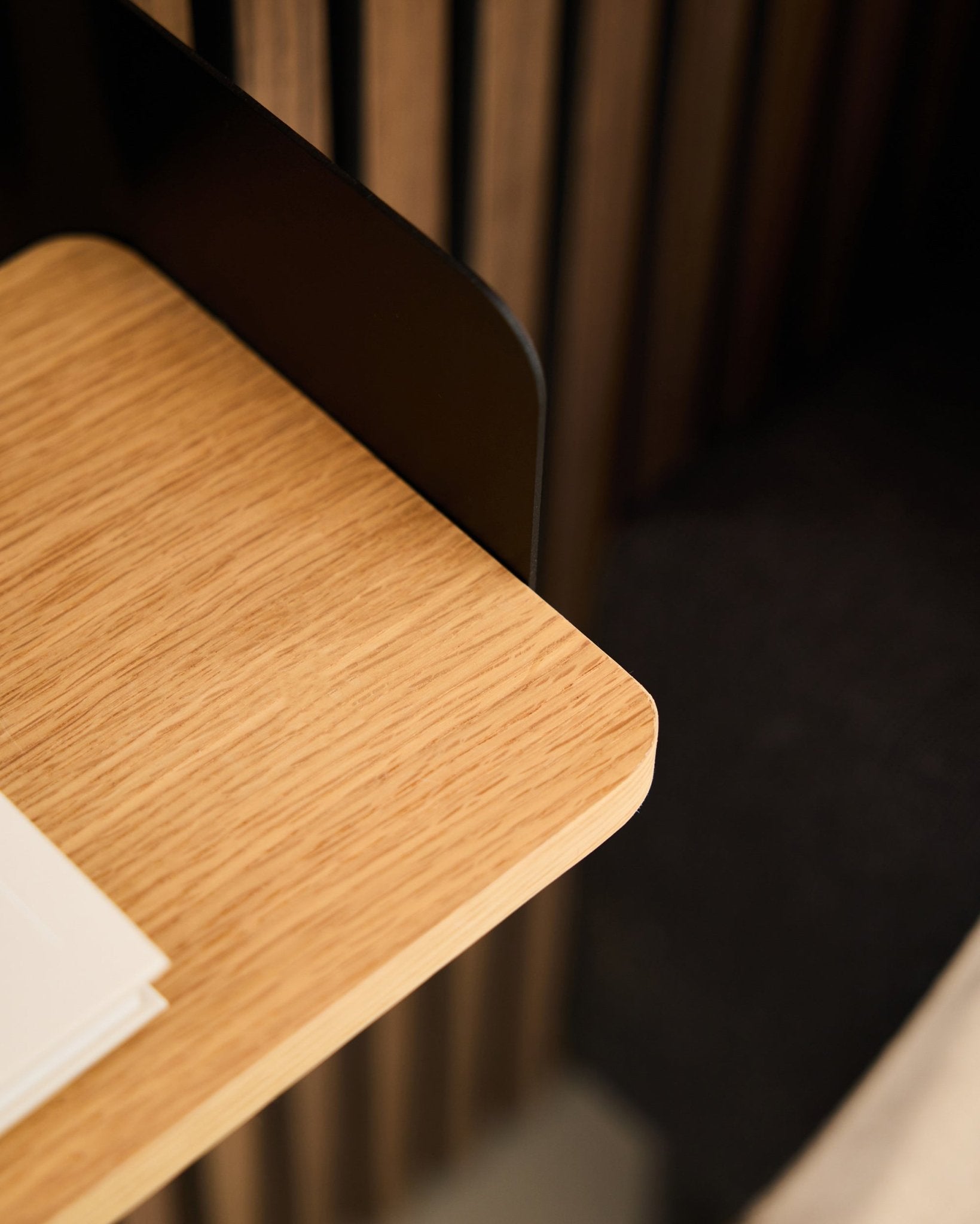 Close-up of a desk corner with black Akupanel acoustic wood paneling, white papers, and the WoodUpp Linn Bookshelf in Natural Oak (39.9 cm) in the background.