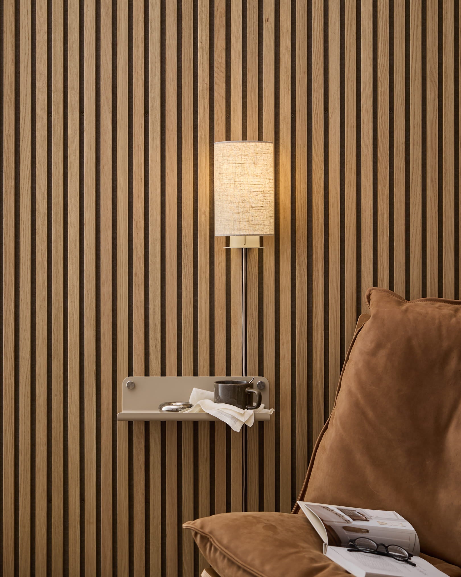 A beige linen-cotton WoodUpp Feya Wall Lamp mounted above a cozy armchair and side table with book and cup, set against a Nordic wood slat acoustic panel.