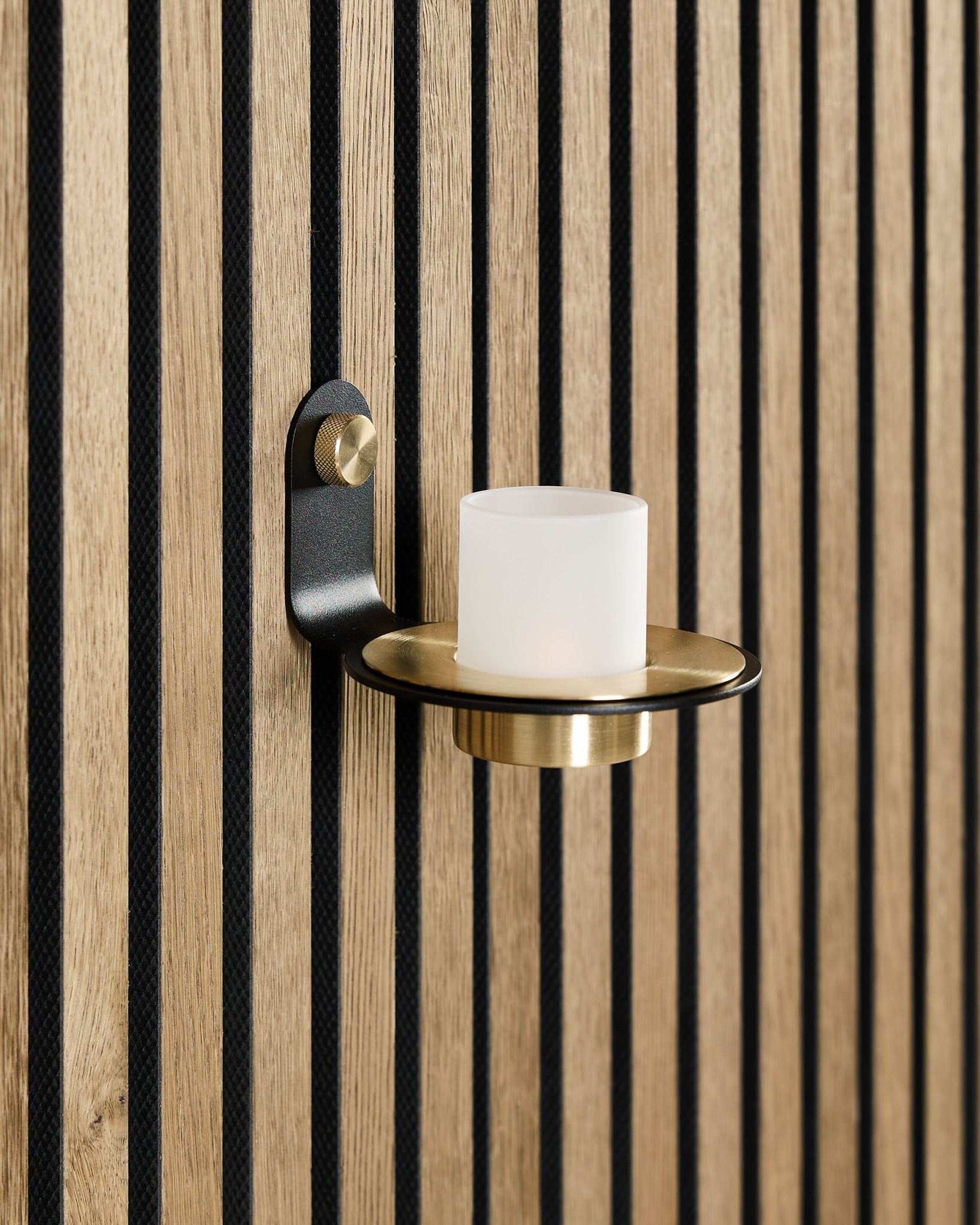 The Norr Candle Holder in black and brass by WoodUpp is mounted on a Nordic Akupanel acoustic wood panel with vertical black slats.