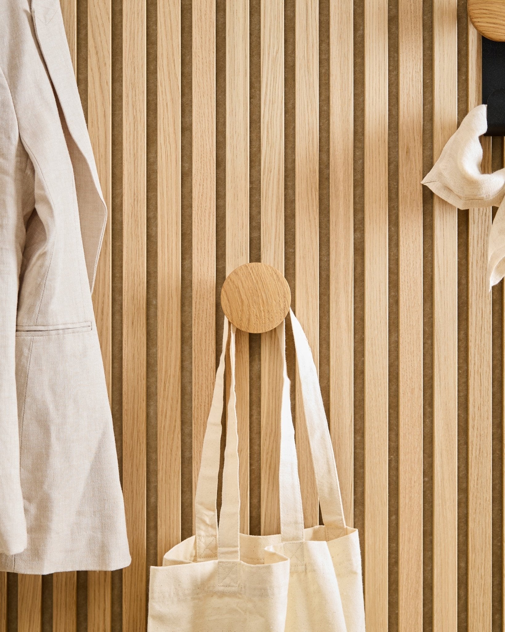 A beige coat and tote bag hang on the Linn I Single Hanger in Natural Oak by WoodUpp, mounted on a vertical Akupanel acoustic wood slat panel.