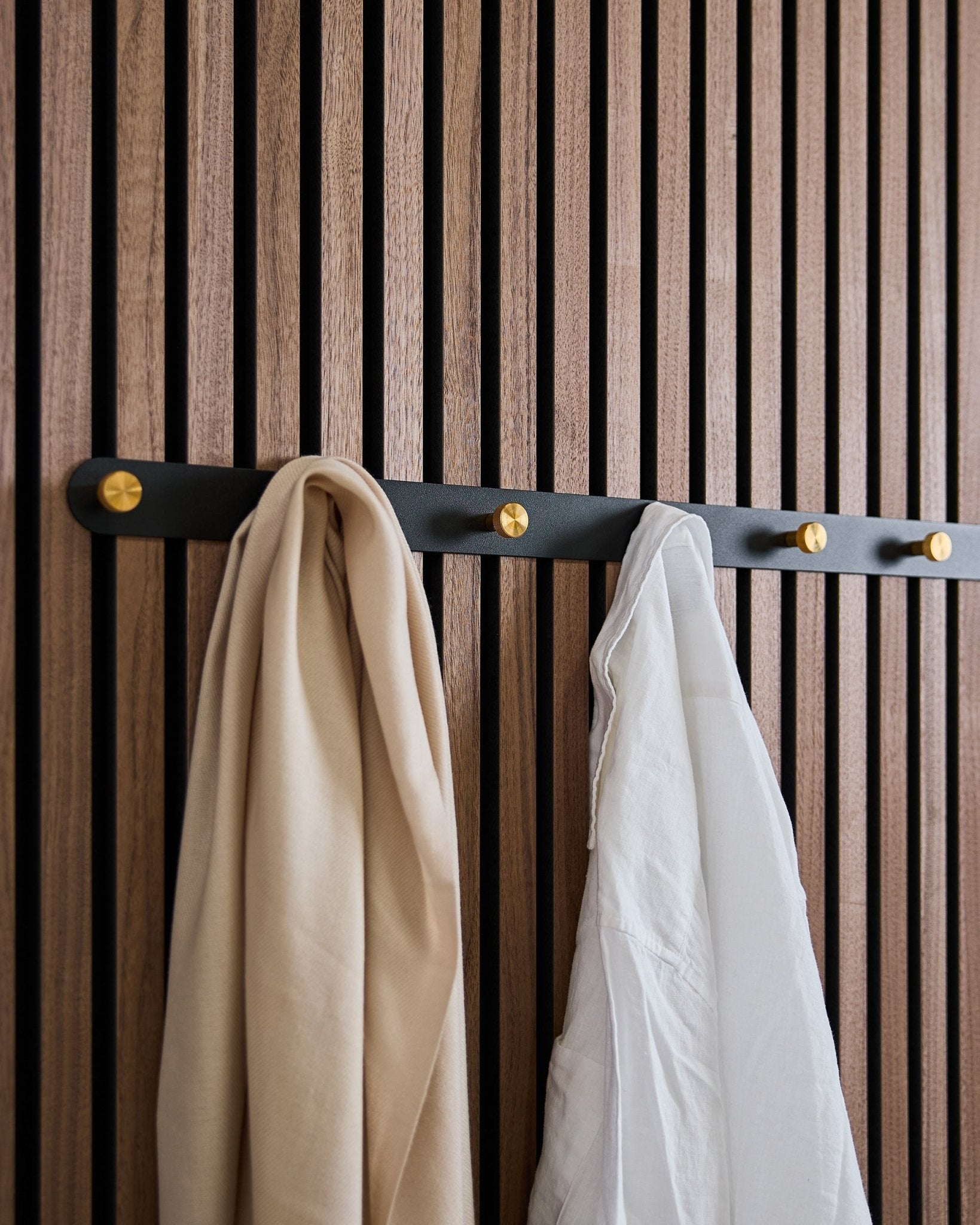 The WoodUpp Norr Coat Hook Rack in black/brass (X7) with a beige scarf and white shirt hangs on a Nordic wooden acoustic slatwall panel.