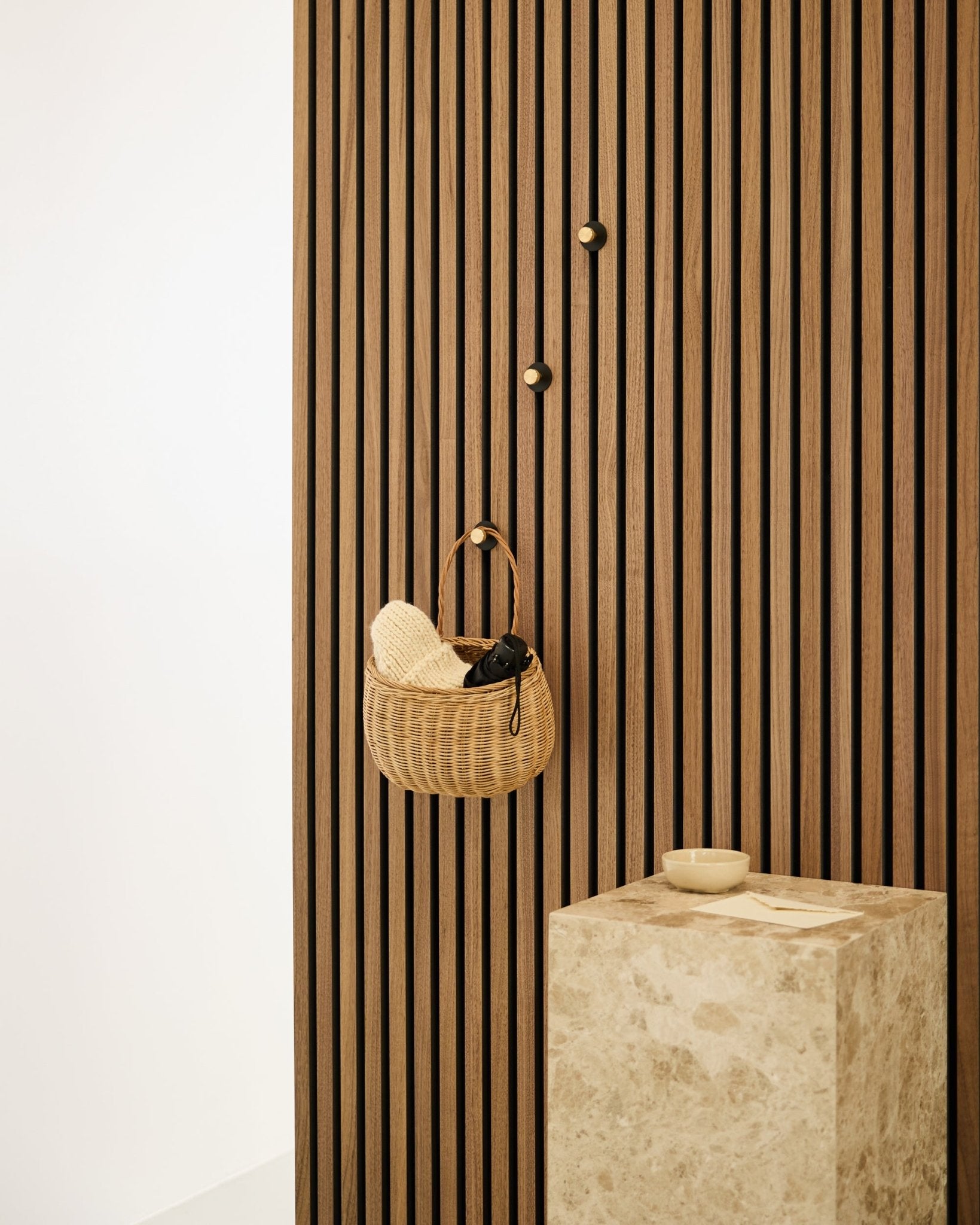 Norr Coat Hook by WoodUpp mounted on a Nordic Akupanel acoustic slat wall, next to a beige stone pedestal with a small bowl in a minimalist interior.
