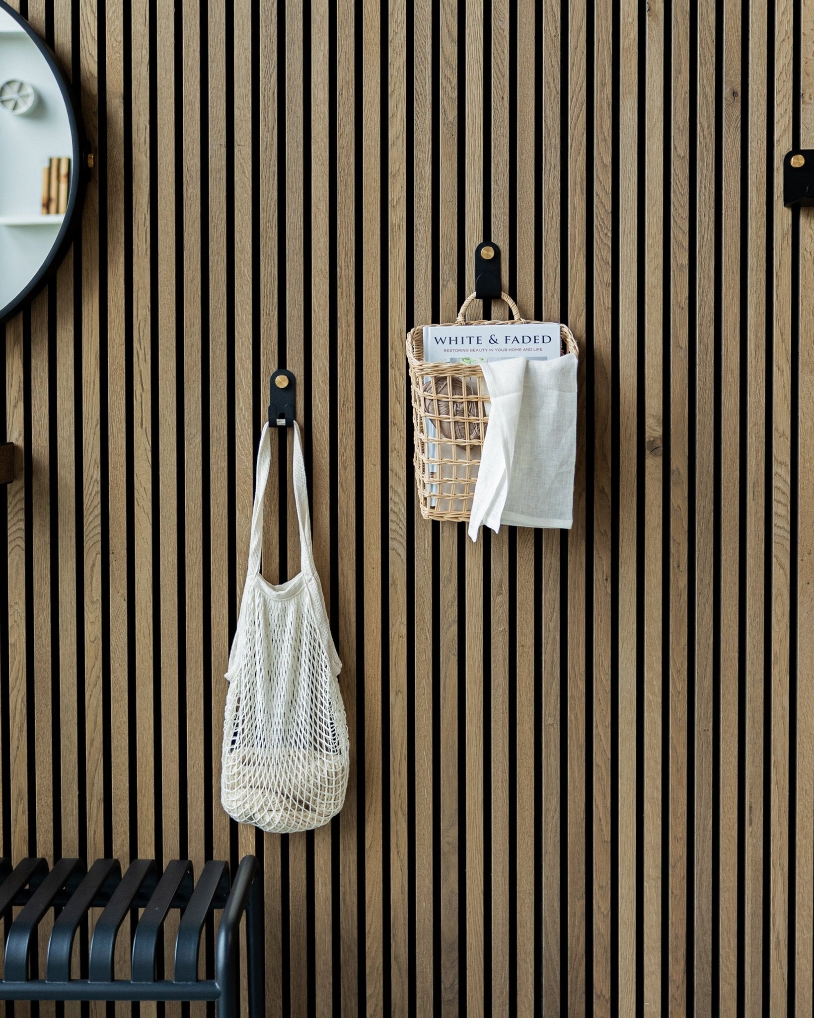 A mesh bag and basket hang from Norr Single Hangers by WoodUpp on a Nordic wooden Akupanel acoustic panel; part of a mirror is visible on the left.