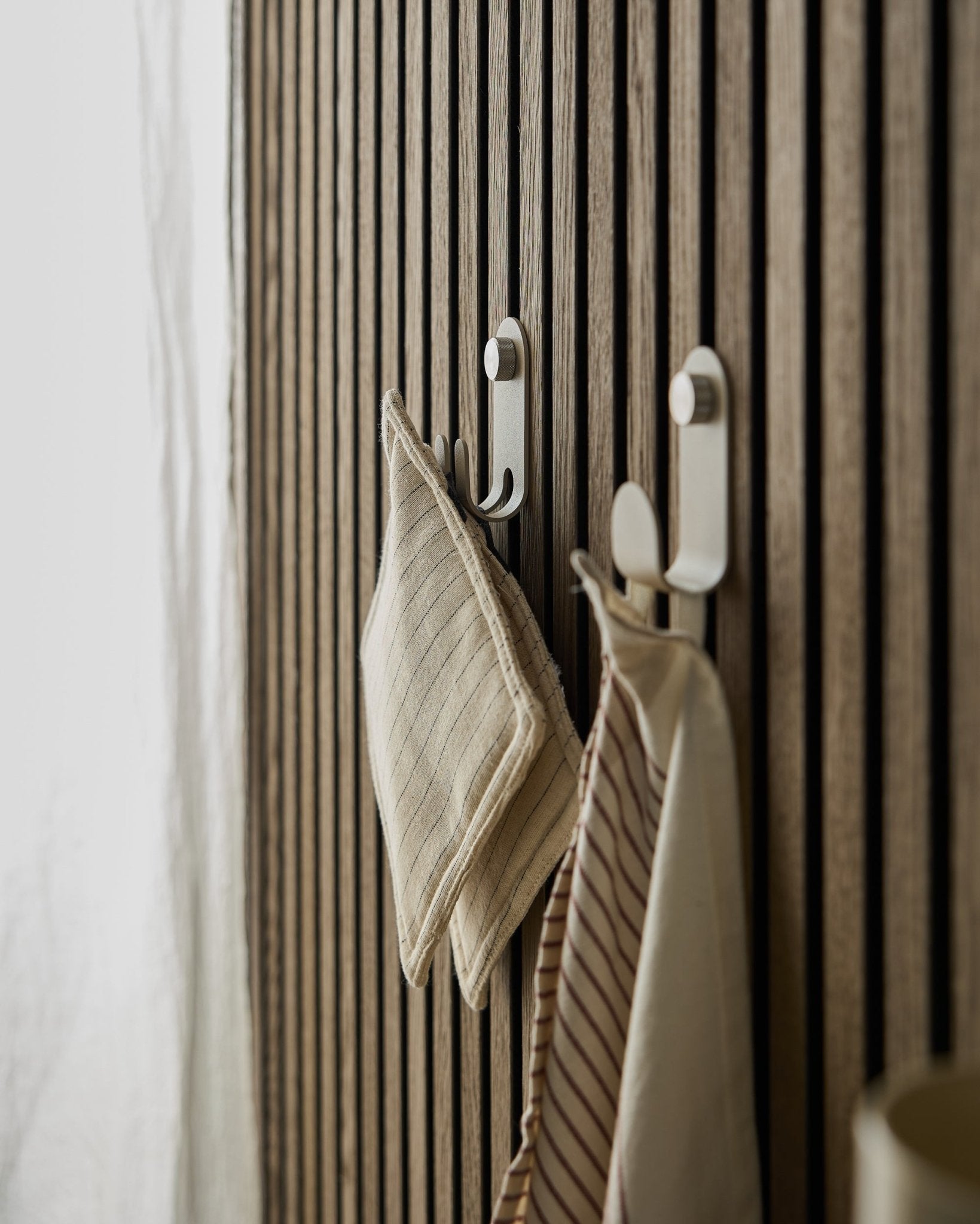 Two striped kitchen towels hang on the Norr Double Hanger by WoodUpp, mounted on a beige Nordic slatwall acoustic panel, illuminated by soft natural light.