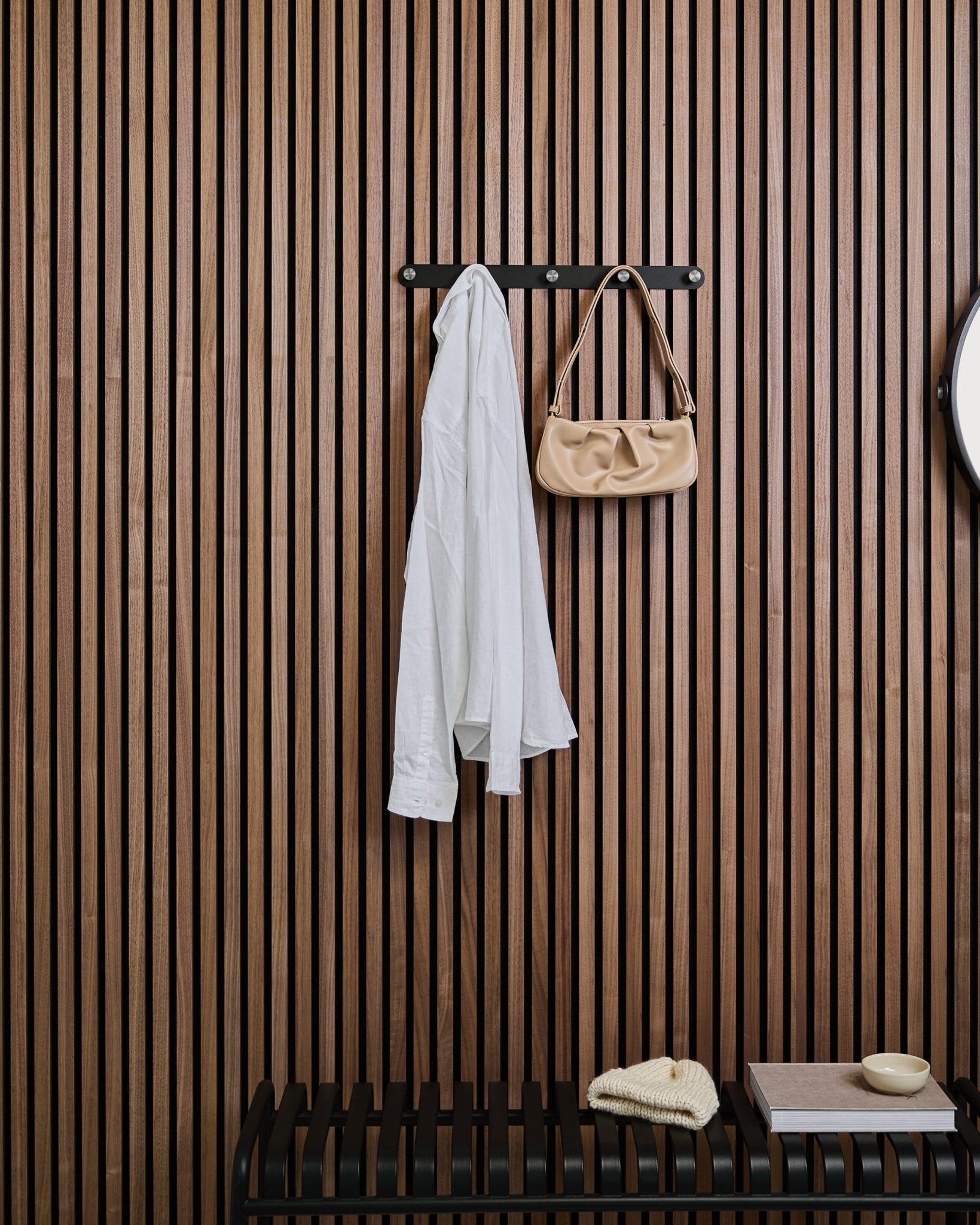 A white shirt and beige handbag hang on WoodUpps Norr Coat Hook Rack above a bench, set against a wooden Akupanel slatwall panel with decor items.