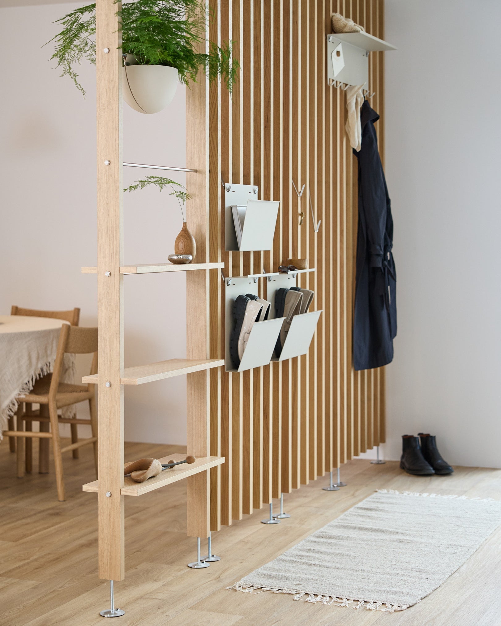 WoodUpp Nell Magazine Storage in beige integrated into a room divider with steel shelves, storage bins, and coat hooks; dining area and rug visible behind.
