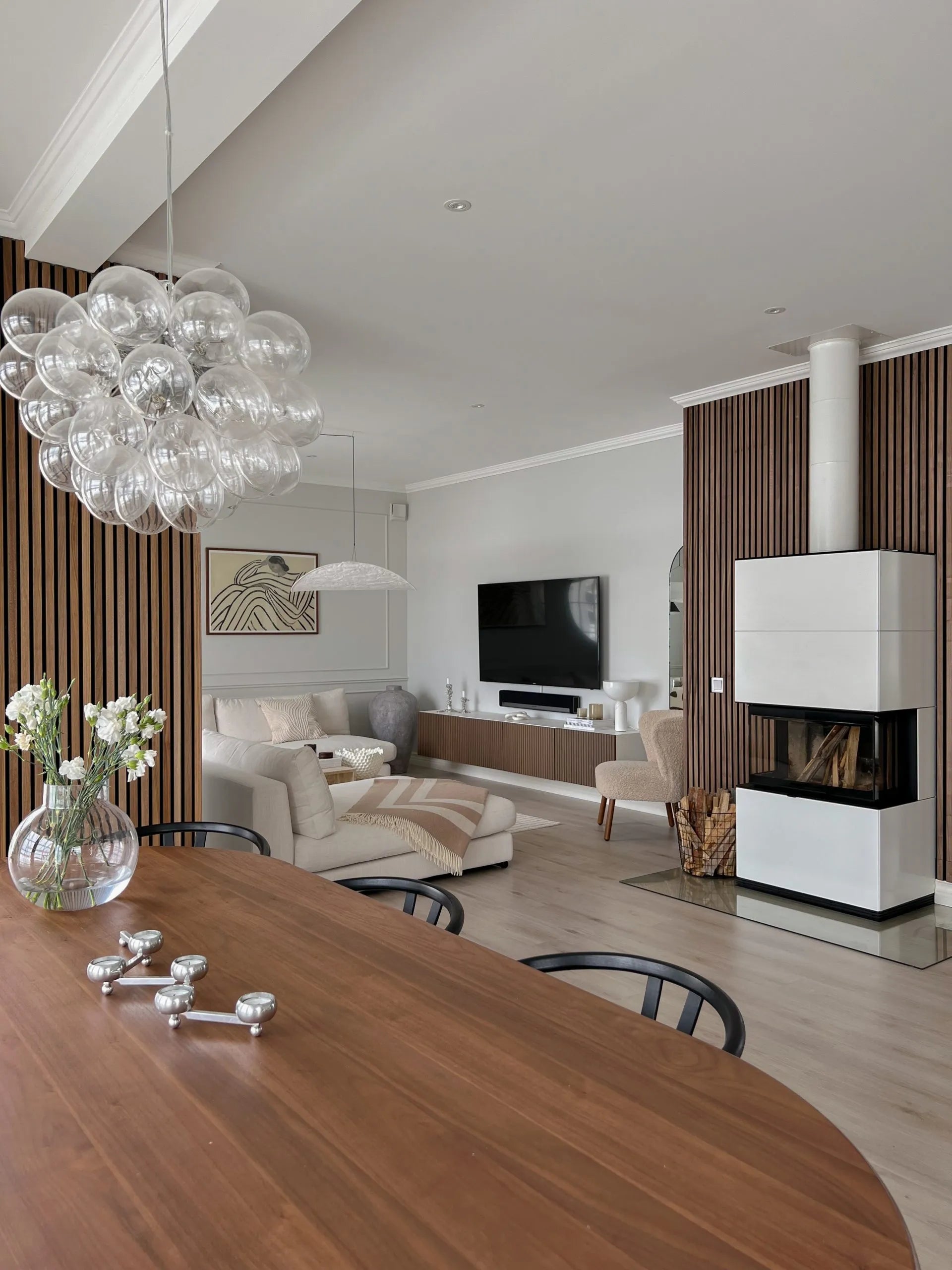 Modern living and dining area featuring Nordic wood acoustic panels, cozy seating, TV, sleek fireplace with stacked firewood, and a glass bubble chandelier.