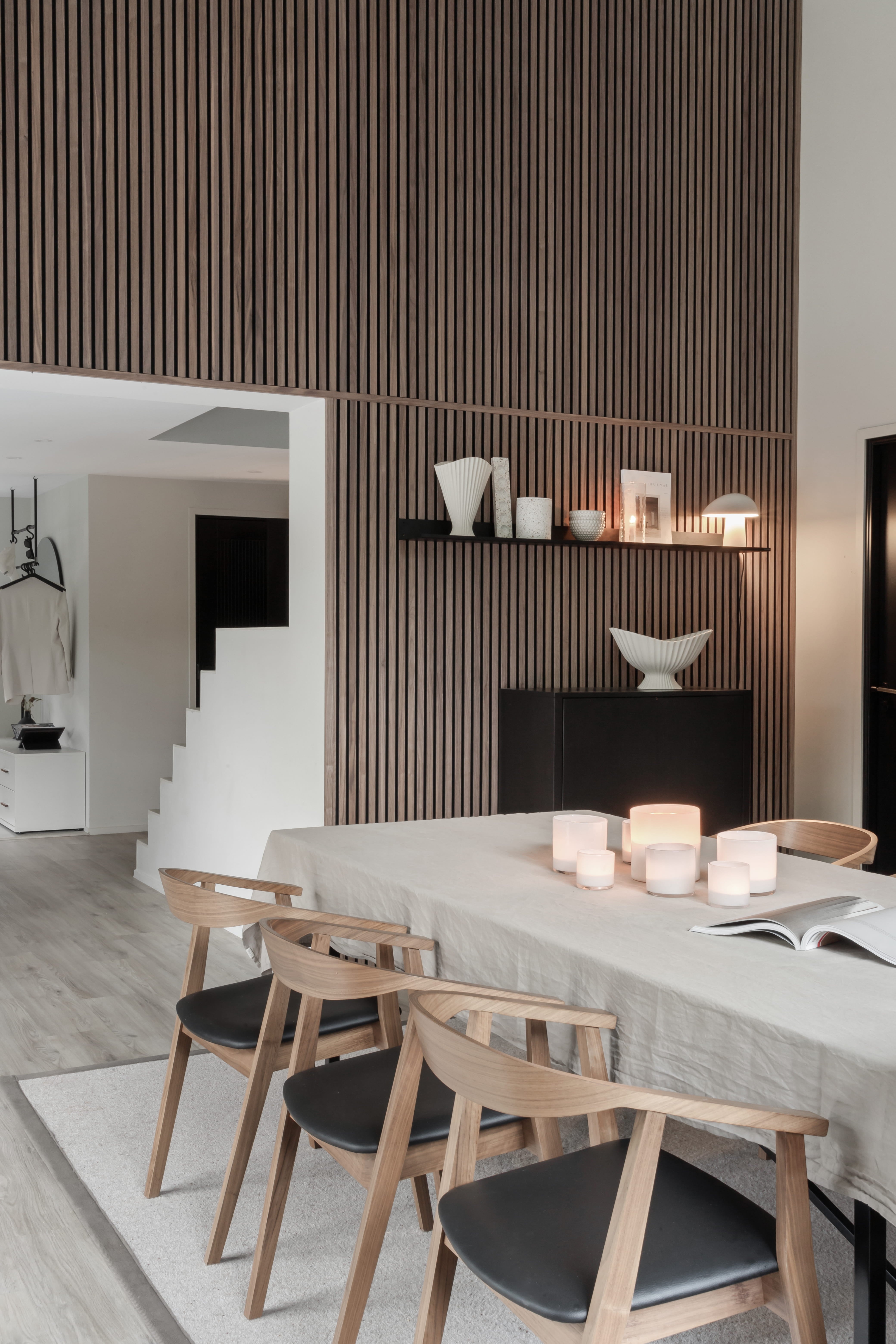 Modern dining room with a light wood table, four chairs, candles, and minimalist decor. Vertical Akupanel acoustic wood slat wall and open staircase in view.