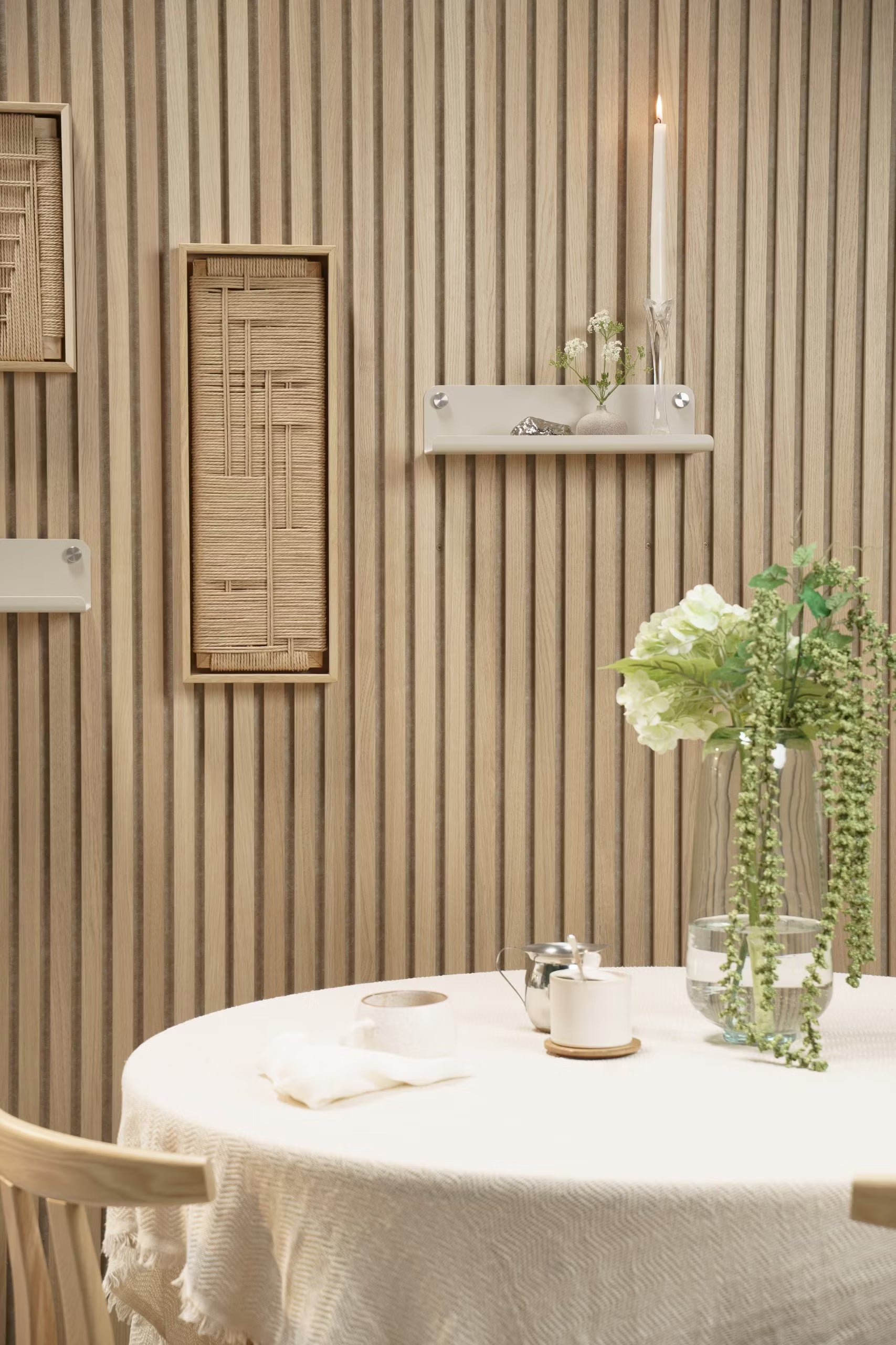 A round table with a light tablecloth, fresh flowers in a vase, and a candle sits in front of a Nordic-style Akupanel acoustic wood slat wall with minimal shelves.