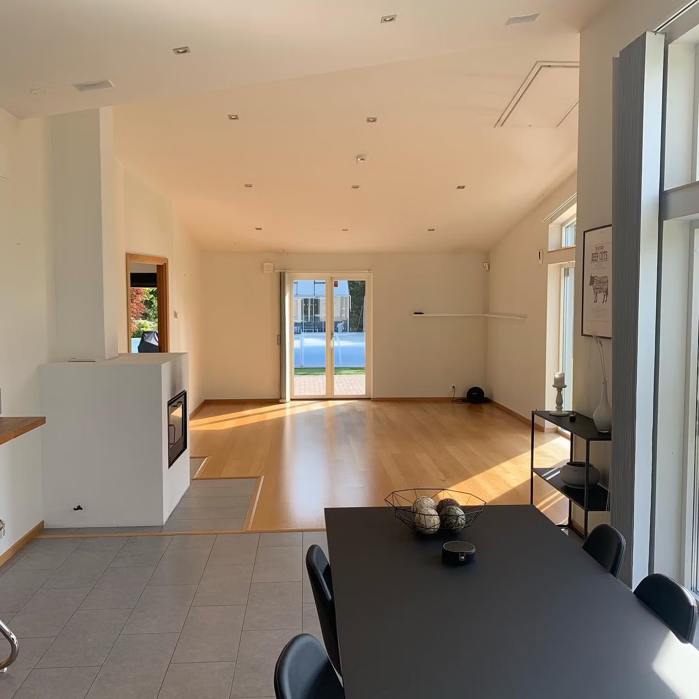 Open-plan room with Nordic wooden floors, Akupanel acoustic wood panels on the wall, large windows, black dining table; doors open to pool and sunny outdoor area.