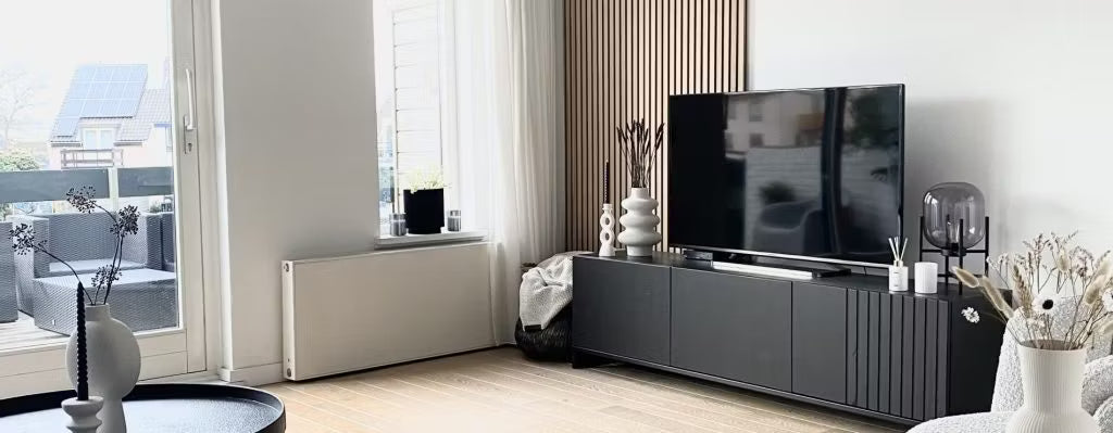 Modern living room featuring a black TV stand, flat-screen TV, decorative vases, and large windows with natural light overlooking the patio; Akupanel on one wall.