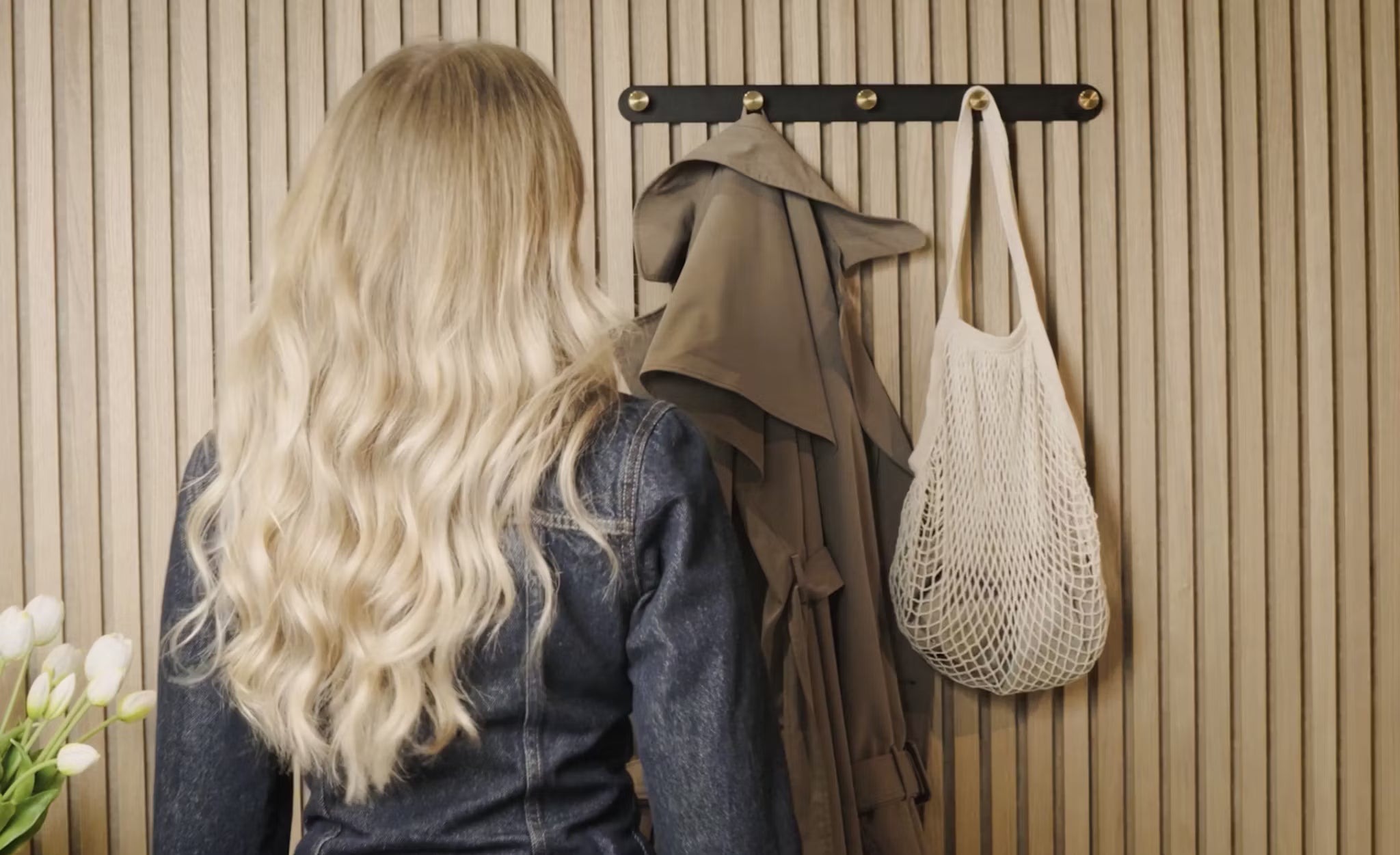 A woman with long blonde hair stands facing a coat rack with a trench coat, white net bag, and fresh white tulips beside an Akupanel acoustic wood panel in the corner.