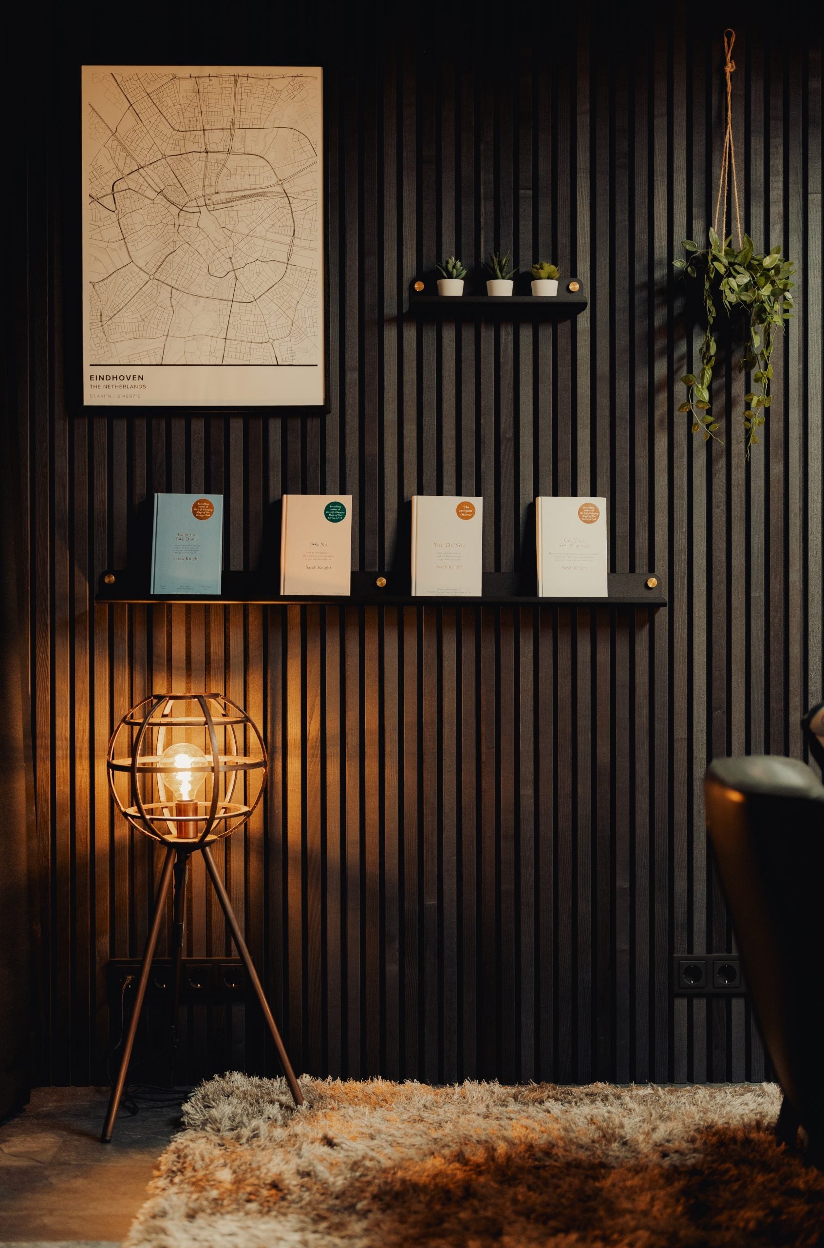 Modern Nordic room with glowing lamp, shag rug, and wall shelves holding books, small plants, and an acoustic panel featuring a framed travel map above.