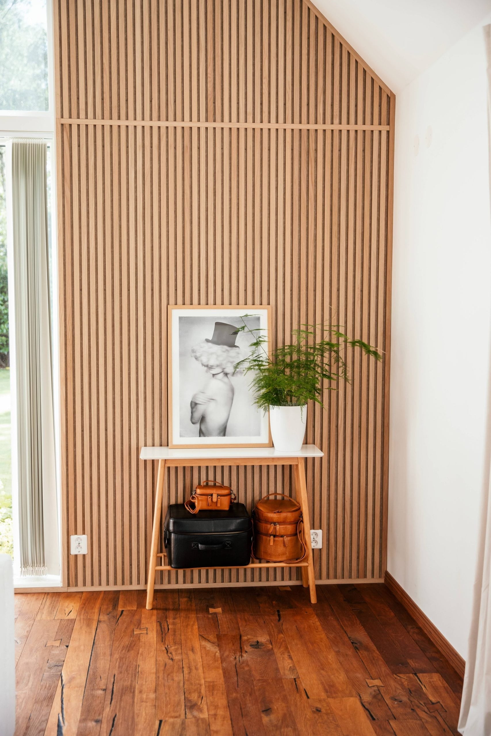 A small wooden table with bags, a framed photo, and a potted plant stands against a Nordic acoustic slatwall panel in a bright room with wooden floors.