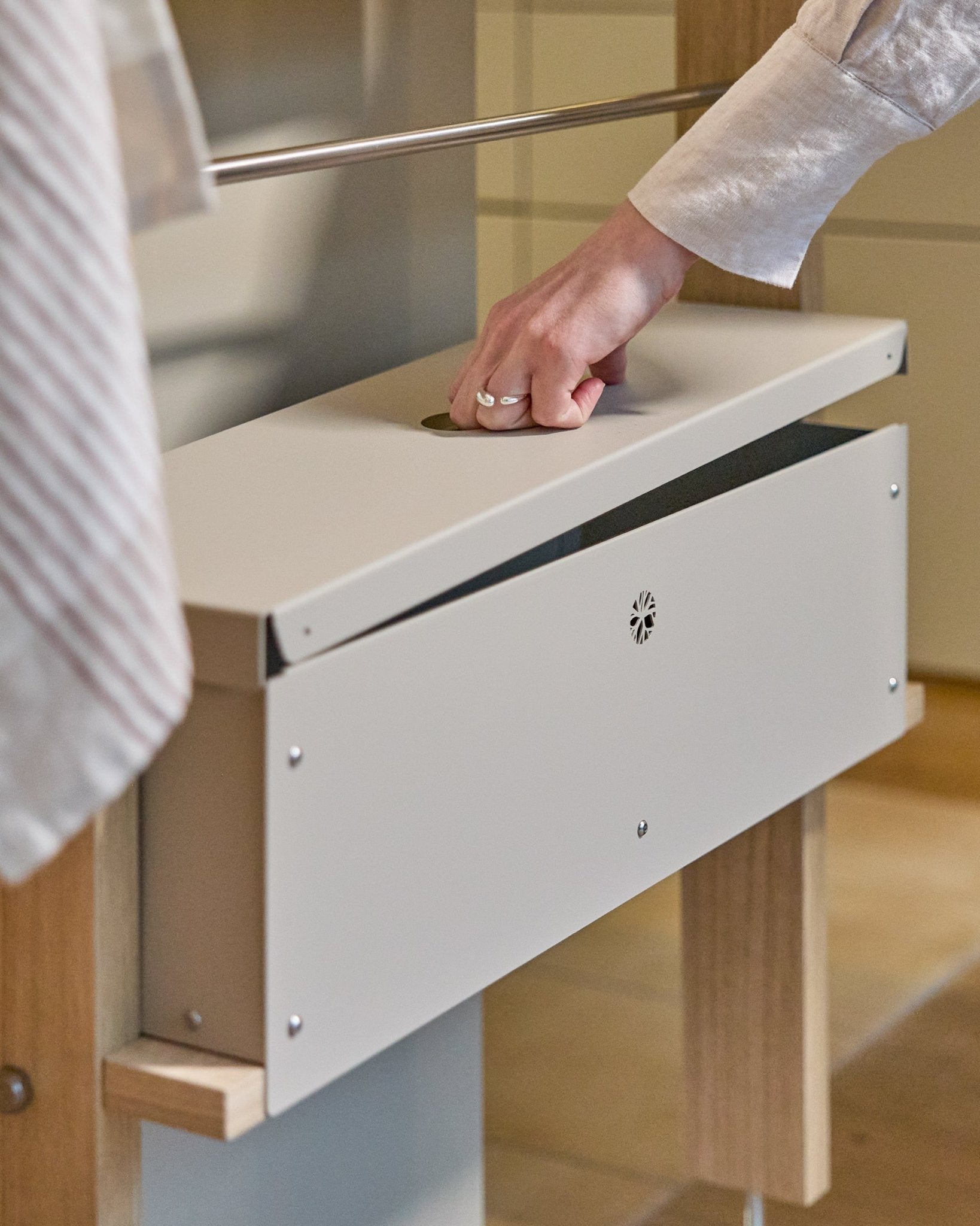 A hand lifts the lid of the WoodUpp Nell Metal Box BM2 in beige, featuring powder-coated steel and wooden supports, designed for organized Nordic storage.