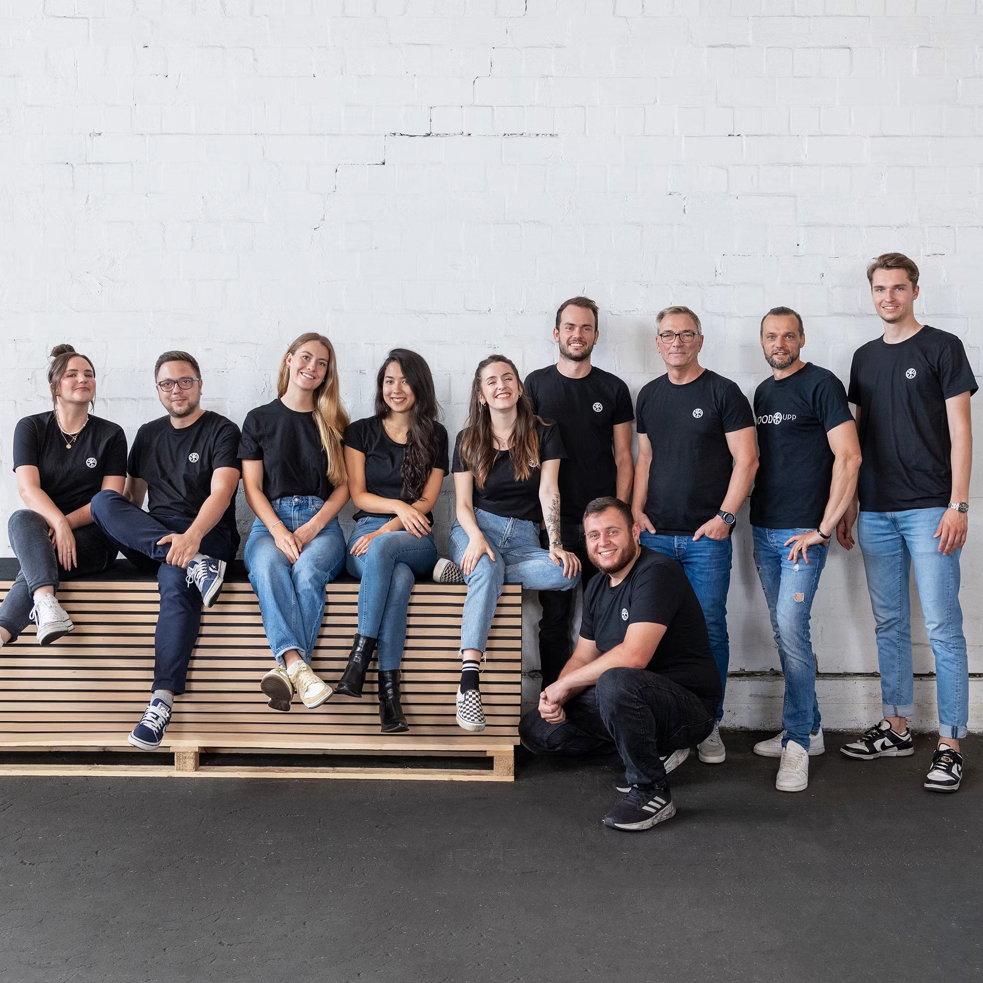 Ten people in matching black t-shirts and jeans smile together by a wooden bench, standing in front of a minimalist white brick wall with Nordic design.