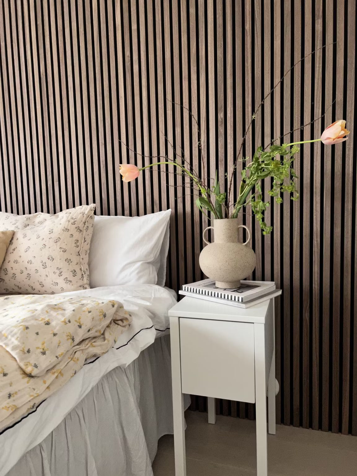 A bedroom with a white bed with floral bedding and a vase of flowers, featuring a Nordic wooden Akupanel acoustic slat accent wall behind the bed.